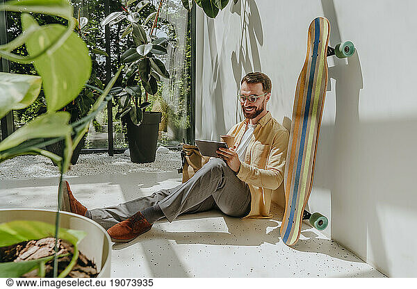 Happy businessman sitting by longboard and using tablet PC in office