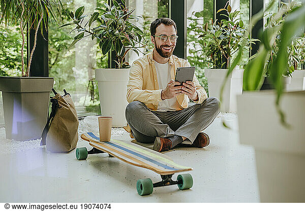 Happy businessman sitting with longboard and using tablet PC amidst plants in office