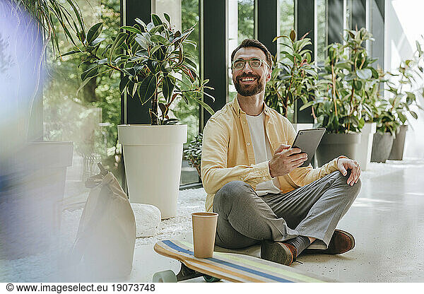 Happy businessman sitting with tablet PC on floor in office