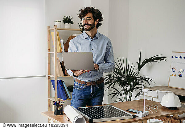 Happy businessman standing with laptop by desk in office