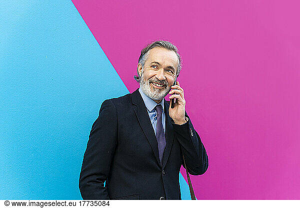 Happy businessman talking on smart phone standing in front of pink and blue wall