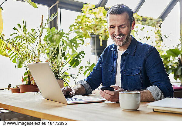 Happy businessman text messaging through smart phone at desk in loft office