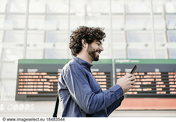 Happy businessman using mobile phone by arrival departure board at station