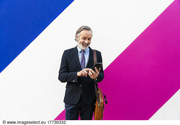 Happy businessman using smart phone standing in front of multi colored wall
