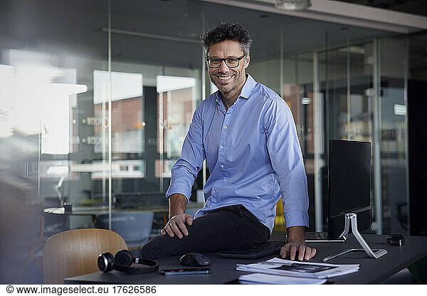 Happy businessman wearing eyeglasses sitting on desk at work place
