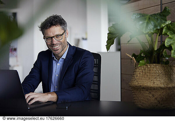 Happy businessman wearing eyeglasses sitting with laptop at desk in office