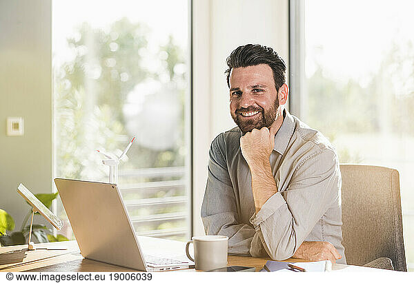 Happy businessman with hand on chin sitting at desk in home office