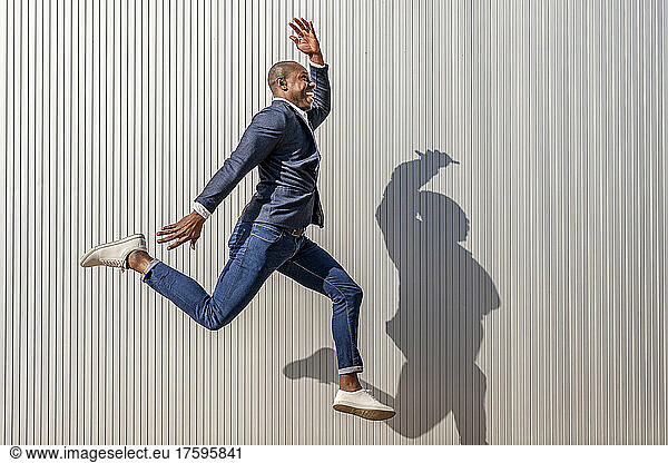 Happy businessman with hand raised jumping by wall on sunny day