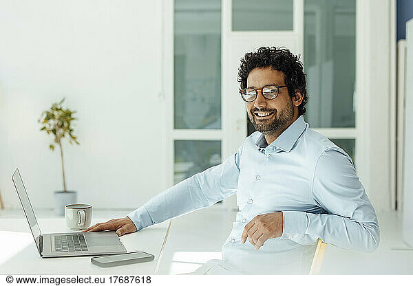 Happy businessman with laptop and mobile phone sitting at desk in office
