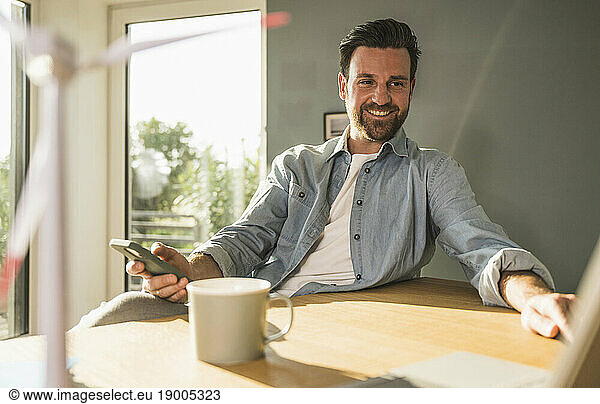 Happy businessman with mobile phone sitting at desk in home office