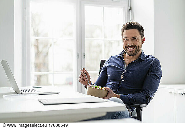 Happy businessman with salad bowl by laptop at office