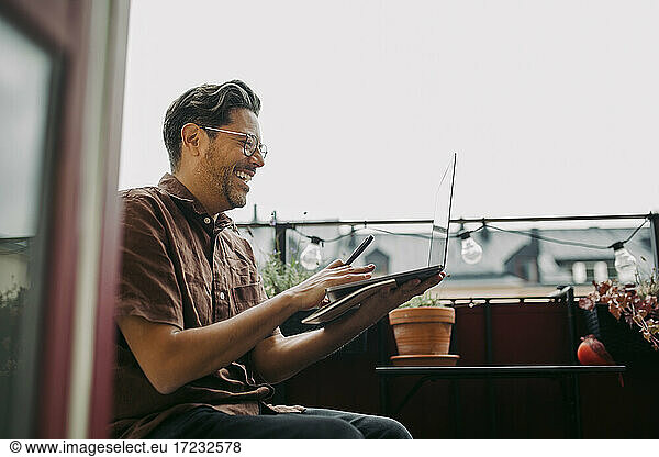 Happy businessman working on laptop while sitting in balcony during COVID-19