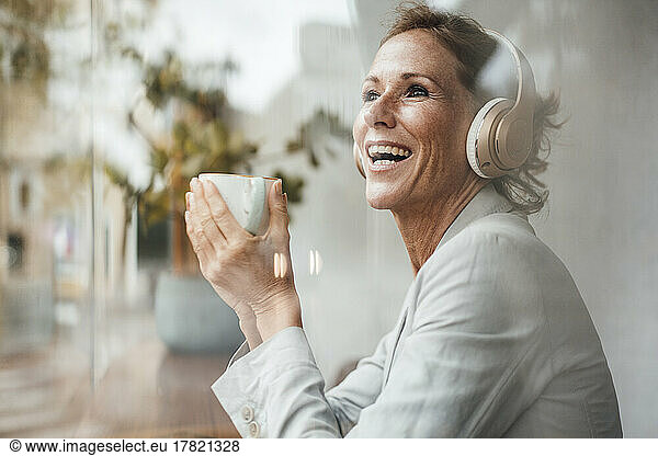 Happy businesswoman holding coffee cup listening music through wireless headphones in cafe