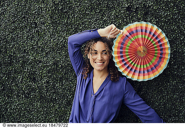Happy businesswoman holding colorful folding fan in front of hedge