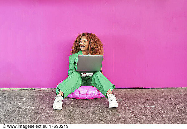Happy businesswoman holding laptop sitting on inflatable ring in front of pink wall