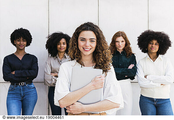 Happy businesswoman holding laptop with colleagues standing in background