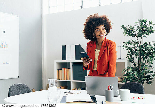 Happy businesswoman holding smart phone standing with laptop at desk in office