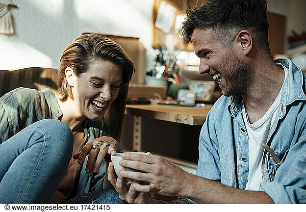 Happy businesswoman looking at cup held by male colleague at home office