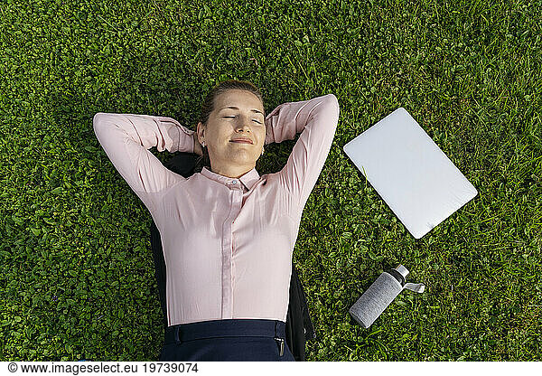 Happy businesswoman lying with laptop and water bottle on grass in park