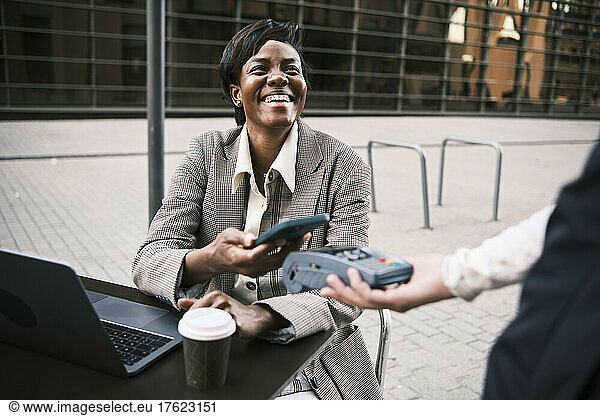 Happy businesswoman making contactless payment through smart phone to waitress at sidewalk cafe