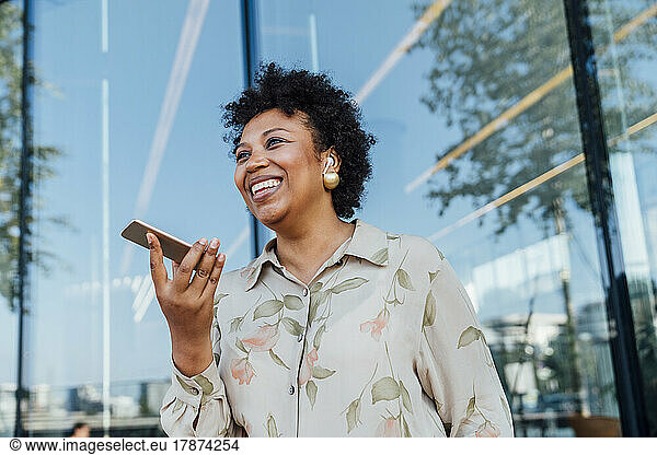 Happy businesswoman sending voicemail through smart phone in front of glass wall