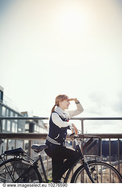 Happy businesswoman shielding eyes while standing with bicycle on bridge against sky