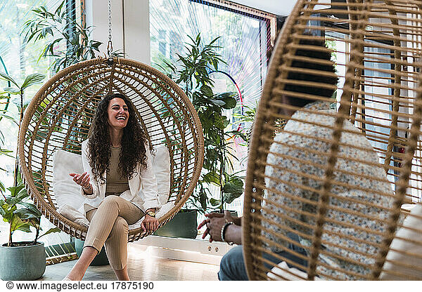 Happy businesswoman sitting in hanging chair talking to colleague