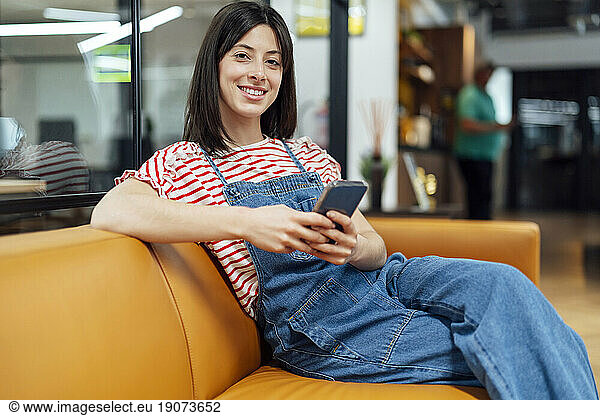 Happy businesswoman sitting with smart phone on sofa in office