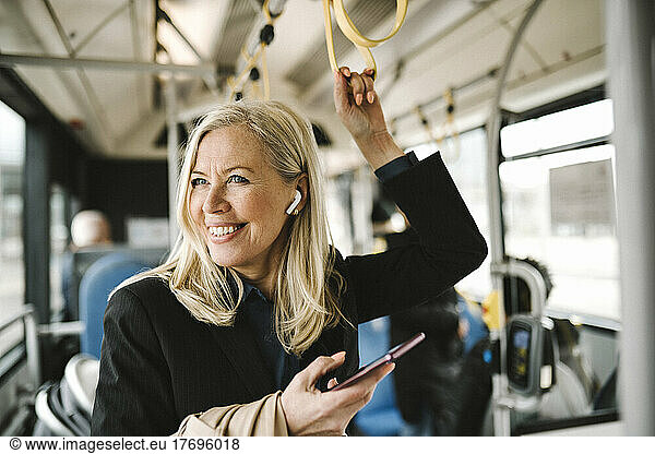 Happy businesswoman talking on smart phone holding grab handle while commuting through bus