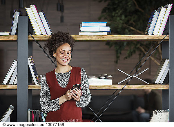 Happy businesswoman using smart phone while standing against book shelf