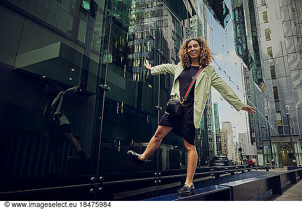 Happy businesswoman with arms outstretched balancing on bench near office building