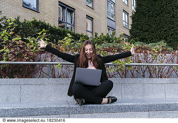 Happy businesswoman with arms outstretched with laptop sitting on bench