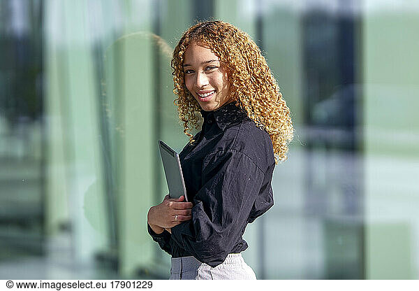 Happy businesswoman with curly hair standing in front of glass