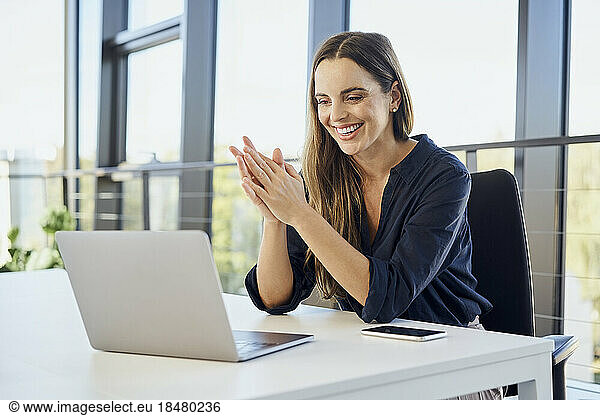 Happy businesswoman with hands clasped doing video call on laptop at office