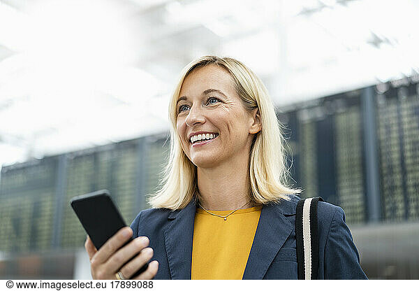 Happy businesswoman with mobile phone in front of arrival board