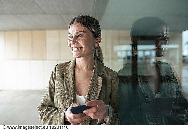 Happy businesswoman with smart phone leaning on glass wall