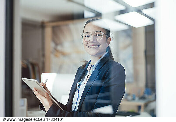 Happy businesswoman with tablet PC seen through glass of office