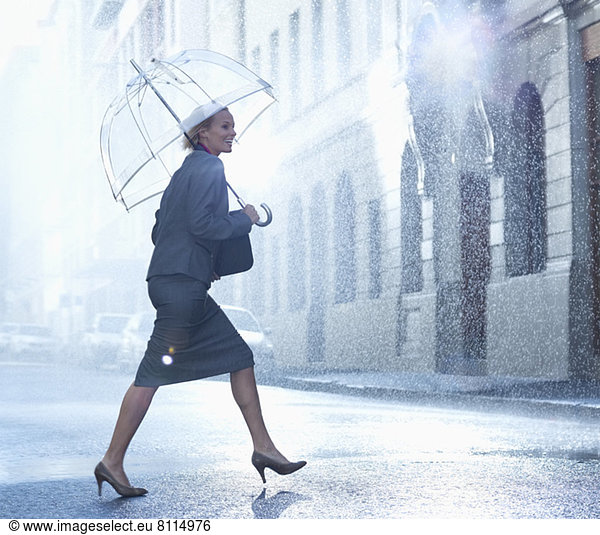 Happy businesswoman with umbrella walking across rainy street