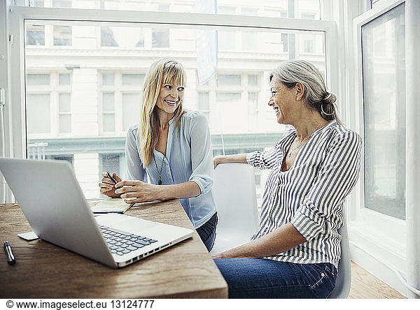 Happy businesswomen talking while sitting at desk in creative office