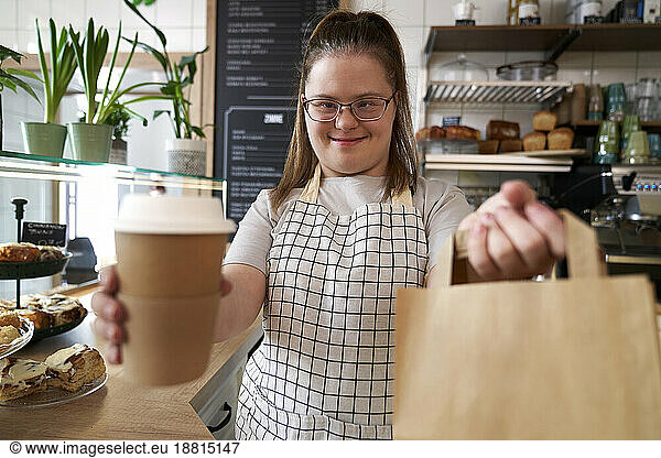 Happy cafe owner with down syndrome holding paper bag and disposable coffee cup in coffee shop