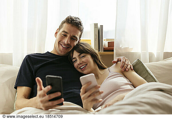 Happy couple with smart phones relaxing on bed at home