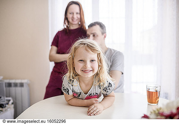 Happy cute girl leaning on dining table with parents standing in background at home