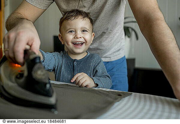 Happy cute son helping father to iron clothes at home