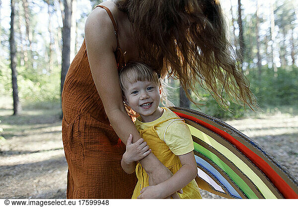 Happy daughter wearing artificial wings standing with mother in forest