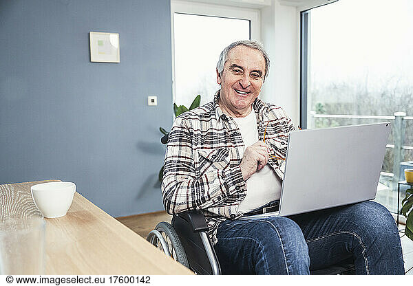 Happy disabled man with laptop sitting on wheelchair at home