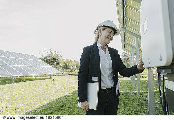 Happy engineer standing with laptop and examining solar panels