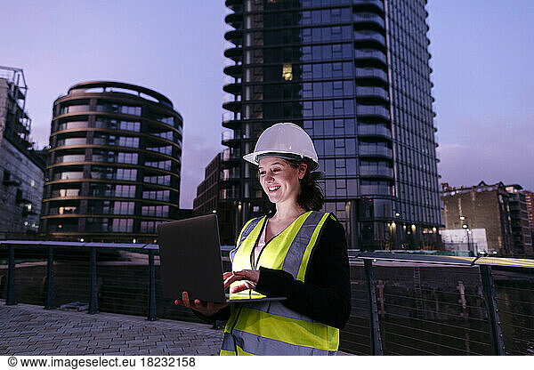 Happy engineer using laptop in front of building at dusk