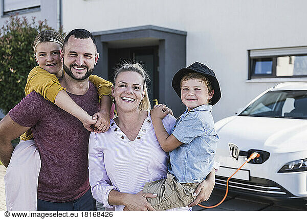 Happy family standing in front of family home and electric car