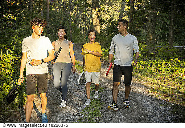 Happy family with tennis ball and rackets walking on dirt road in forest during vacation