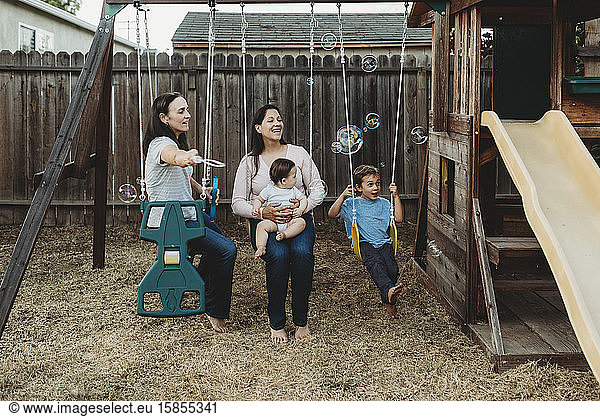 Happy family with two moms on swings playing with bubbles